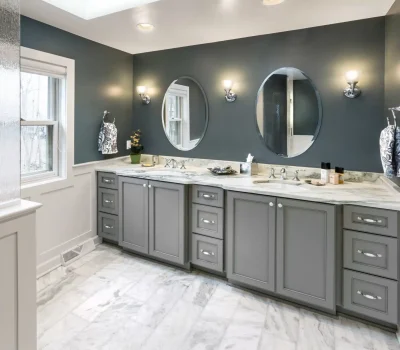 Modern bathroom with gray cabinets, marble countertops, and two oval mirrors. Soft lighting and decorative towels accentuate the sophisticated design and style.