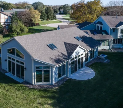 Suburban neighborhood with modern house, large windows, and skylights. Surrounding greenery includes trees and lawns. Quiet, winding road in the background.