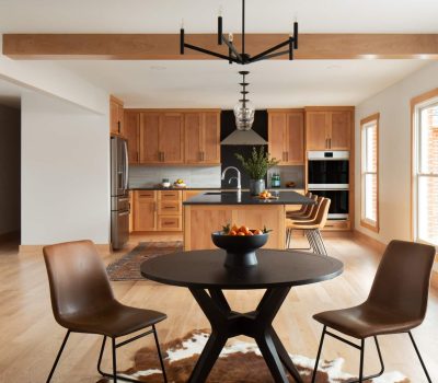 Modern kitchen with wooden cabinets, island with stools, round dining table with chairs, and fruit bowls. Bright, spacious layout with natural light.