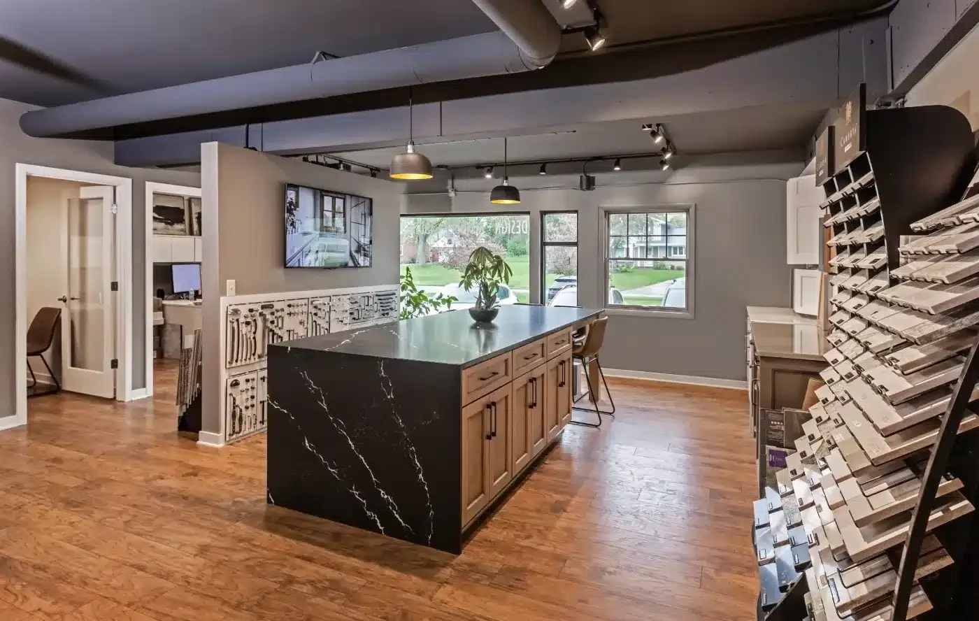Modern interior design studio with large central island, wooden flooring, architectural samples, and window view. Open space features desks, seating, and decor.