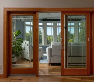 A cozy interior features large windows, light gray sofas, a potted plant, and wooden sliding doors with a view of lush green trees.