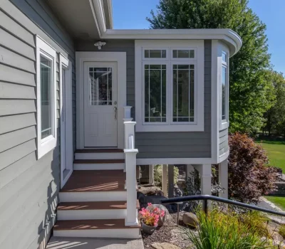 A gray house features a small porch, white door, and windows. The lush garden includes colorful flowers and green shrubs under a clear blue sky.