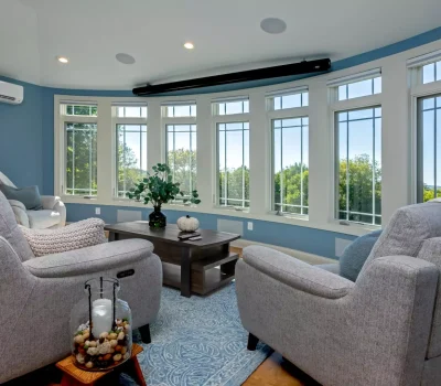 Bright living room with large windows, gray sofas, and a wooden table. Blue walls create a calming atmosphere, overlooking a lush, green landscape.