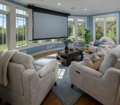 Cozy living room with light gray sofas, a dark wood coffee table, projector screen, large windows, blue walls, and lush outdoor view.