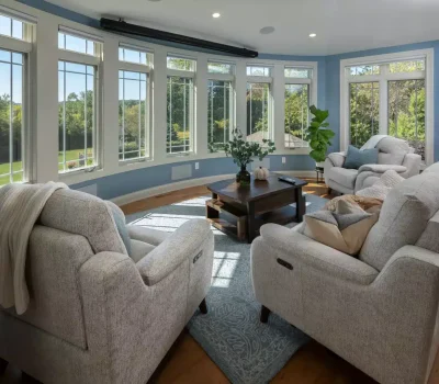 Bright, cozy living room with large windows, grey sofas, and wooden coffee table set against blue walls, overlooking a lush green outdoor view.