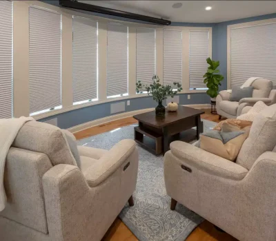 A cozy living room with blue walls, beige furniture, a wooden coffee table, and plants. Window blinds cover large, curved windows.