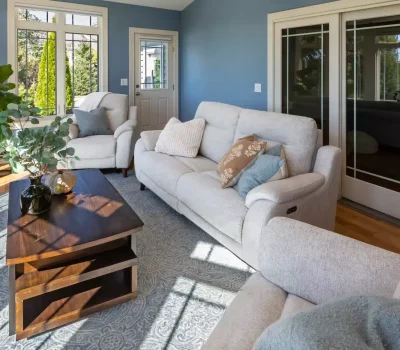 Bright living room with blue walls, white sofas, large windows, and a wooden coffee table with a plant. Sunlight streams in through windows.
