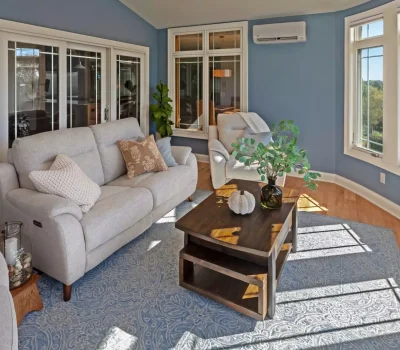 Bright living room with blue walls, white sofas, a wooden coffee table, and large scenic windows. Decor includes plants and cozy cushions.