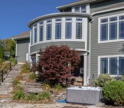 Modern house with large windows, stone steps, and landscaping. Features a hot tub and basketball hoop in the backyard under a clear blue sky.