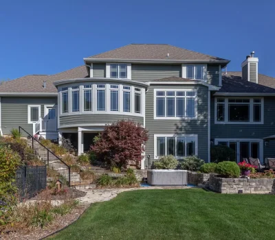 A large modern house with abundant windows overlooks a lush garden with stone pathways and patio seating, set under a clear blue sky.