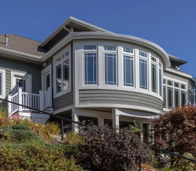 A modern house with large bay windows, surrounded by landscaped greenery and shrubs, against a clear blue sky. No people present.