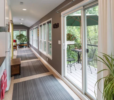 A bright sunroom features large windows, sliding glass doors opening to a deck, plants, a table with chairs, and striped rugs on the floor.