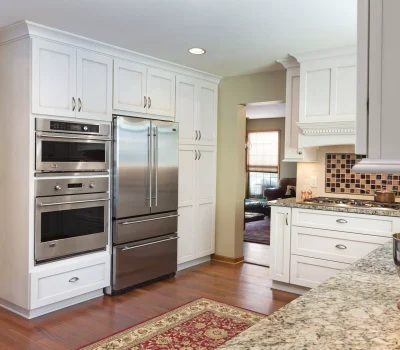 Modern kitchen with granite countertops, stainless steel appliances, and white cabinets. Cozy living room visible through doorway. Hardwood floors and elegant decor.