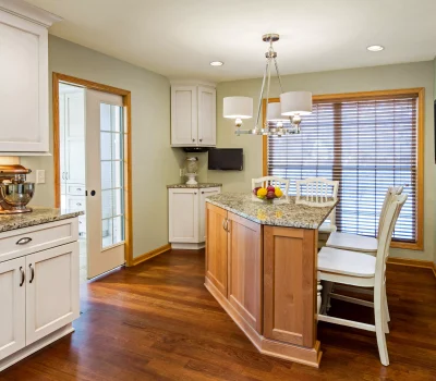 A modern kitchen with wooden floors features a granite island, white cabinets, a chandelier, and a fruit bowl.