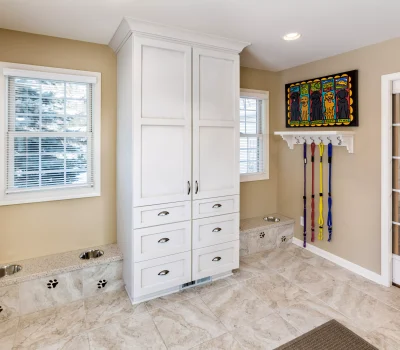 Bright pet-friendly room with wall hooks holding leashes, colorful dog artwork, built-in feeding stations, white cabinetry, and tiled floor.