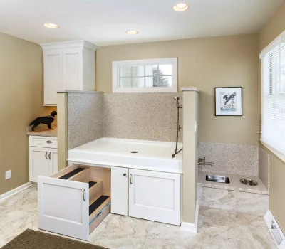 A well-lit room with a dog wash station, pull-out steps, and pet feeding area. Decorated with a dog silhouette and framed artwork.