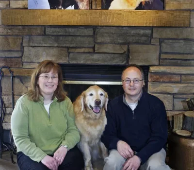 Two persons sitting with a dog in front of a stone fireplace, with dog portraits displayed on the mantel above them.