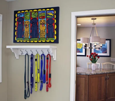 A colorful dog-themed artwork hangs above various leashes. Adjacent, a kitchen features tulips on a countertop under a stylish chandelier.