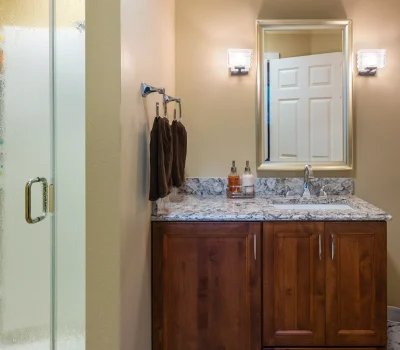 A modern bathroom features a glass shower, wooden vanity with marble countertop, large mirror, wall lights, and neatly hung towels.