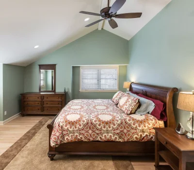 A cozy bedroom with a patterned bedspread, wooden furniture, soft lighting, ceiling fan, green walls, hardwood floor, and window blinds.