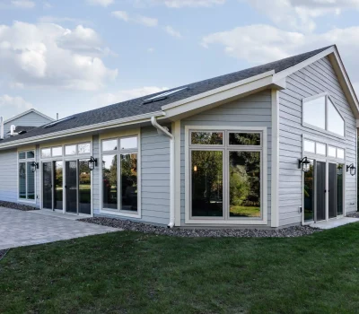 Modern house with large windows, stone patio, and green lawn. Clear blue sky with scattered clouds in the background. Peaceful residential setting.
