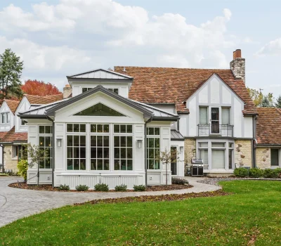 Charming Tudor-style house with white and stone exterior, brown roof, surrounded by trees and manicured lawn. Paved walkway leads to entrance.