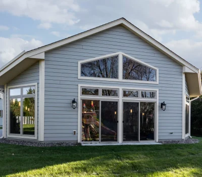 A modern gray house with large windows, set in a grassy yard, surrounded by trees under a partly cloudy sky.