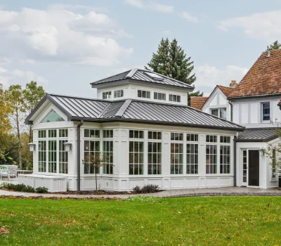 A charming house features a bright sunroom with large windows and a metal roof, surrounded by lush green lawn and trees.