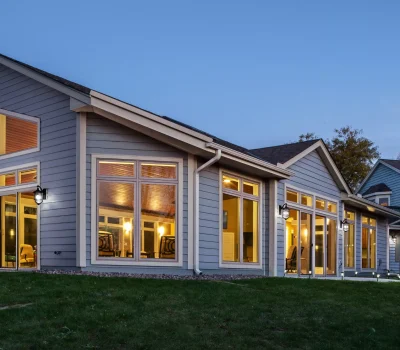 A modern house with large windows illuminated from within during twilight, surrounded by a grassy yard and neighboring homes in a suburban setting.