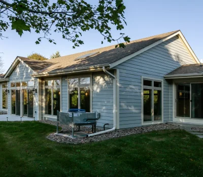 A light gray house with large windows and a well-kept lawn, surrounded by trees under a clear blue sky.