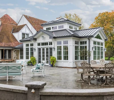 Elegant house with a modern sunroom, surrounded by a spacious patio with chairs, greenery, and a backdrop of autumn trees under a cloudy sky.