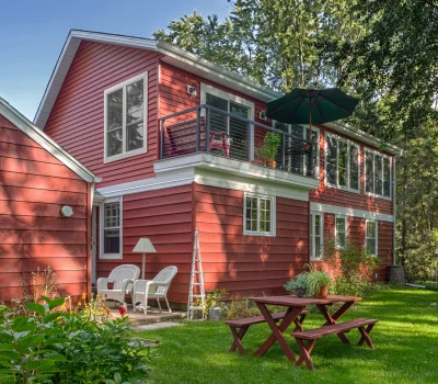 A red two-story house with a balcony, surrounded by trees and a garden, featuring outdoor furniture on the patio and lawn.