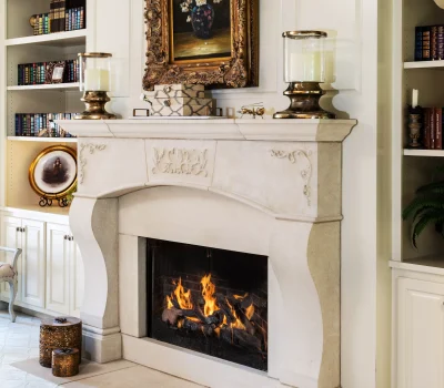 Elegant living room with a stone fireplace, framed floral painting, and surrounding bookshelves filled with books. Cozy flames flicker in the hearth.