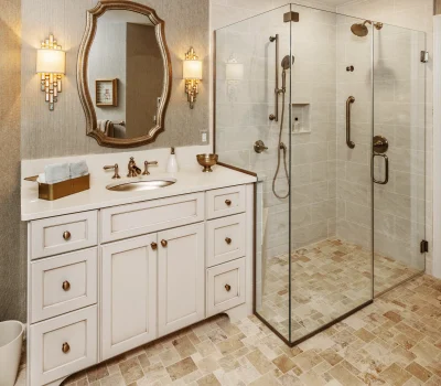 Elegant bathroom with glass shower, beige tiles, and white vanity. Ornate mirror and wall sconces complement the sophisticated, warm design.