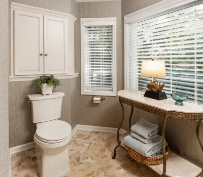 A cozy bathroom with a toilet, cabinet, and decorative table. Lamp and plant add warmth. Natural light enters through two windows.
