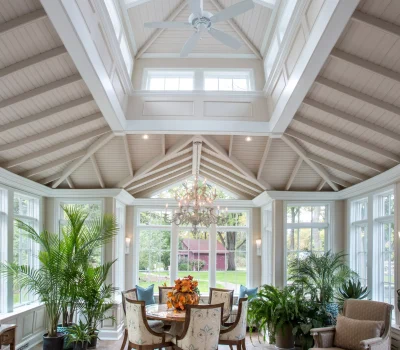 Bright sunroom with vaulted ceiling, chandelier, and fan. Large windows overlook lush garden. Elegant table setting with flowers, surrounded by comfortable chairs and plants.