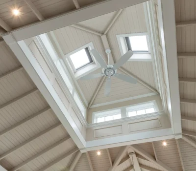 Elegant ceiling with skylights, wooden beams, and a large fan. A chandelier hangs beneath, enhancing the spacious and well-lit interior ambiance.