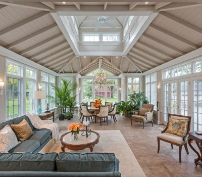 A spacious sunroom with large windows, cozy seating, lush plants, and a chandelier. It's elegantly furnished, creating a bright, inviting atmosphere.