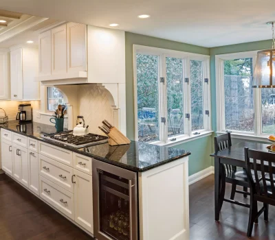 Cozy kitchen with white cabinetry, dark countertops, gas stove, and dining area. Large windows overlook a garden. Warm pendant lighting enhances ambiance.