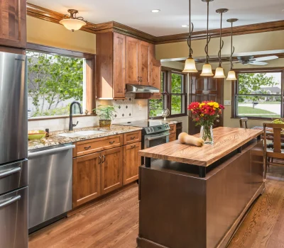 A modern kitchen with wooden cabinets, island, stainless steel appliances, pendant lights, and flowers on the counter. Large windows overlook greenery outside.