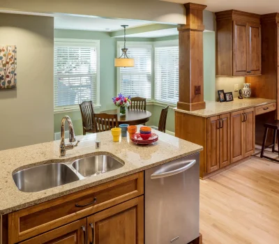 A modern kitchen with granite countertops, a double sink, wooden cabinets, dining area, colorful utensils, and wall art, illuminated by natural light.