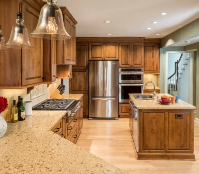 Warm, inviting kitchen with wooden cabinets, stainless steel appliances, pendant lighting, and a central island. Decorative vase with flowers on countertop.