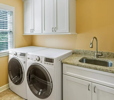 A laundry room with a washing machine, dryer, white cabinets, granite countertop, and a stainless steel sink against a beige wall.