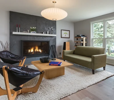 A modern living room with a cozy fireplace, stylish furniture, a Picasso book on a table, and a large window with trees outside.