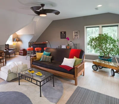 A cozy, modern living room with a black sofa, colorful pillows, glass table, desk, and greenery. Red stool seating near a small bar.