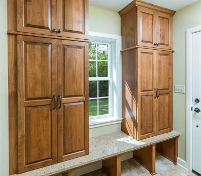 Wooden cabinets and a matching bench line the tiled room, with a window offering a view of greenery outside. Neutral beige walls.