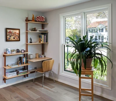 A cozy home workspace with shelves, books, art, a potted plant, and a window view of a white house, surrounded by greenery.