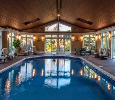 Indoor pool area with wooden ceiling, surrounded by large windows and lounge chairs. Outdoor view through windows shows greenery and playground.