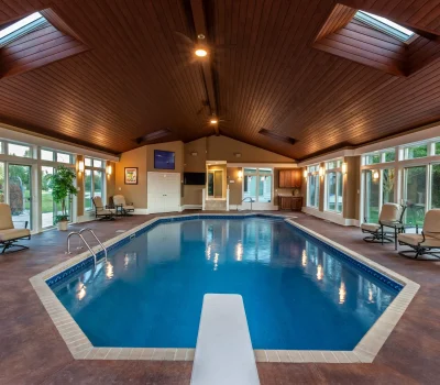 Spacious indoor swimming pool room with skylights and wood ceiling, surrounded by large windows. Seating area with chairs and tables on poolside.