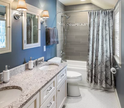 Modern bathroom with dual sinks, granite countertop, blue walls, white cabinetry, and gray tiled shower. Elegant lighting and patterned shower curtain enhance decor.